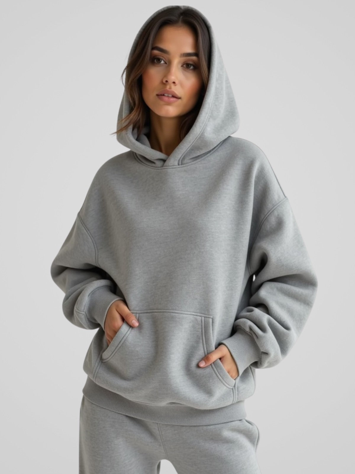 Woman wearing a light gray oversized hoodie, standing and facing forward with hands in the pockets to show the fit and style.